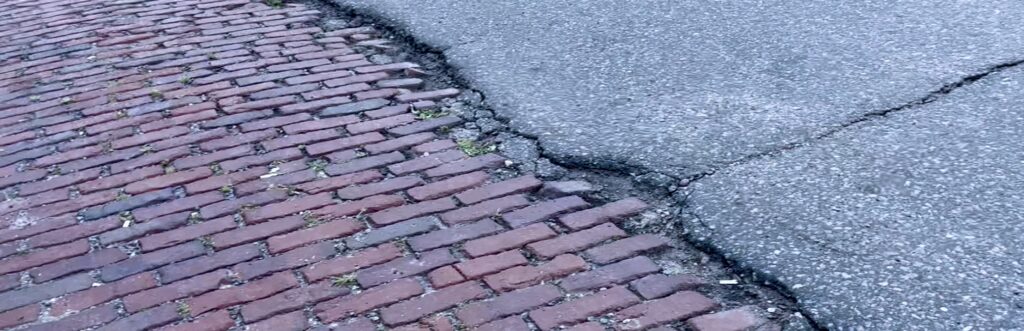 closeup photo of Quincy Street Northeast, with original brick street revealed on the left of the image, partially covered by blacktop on the right side of the image